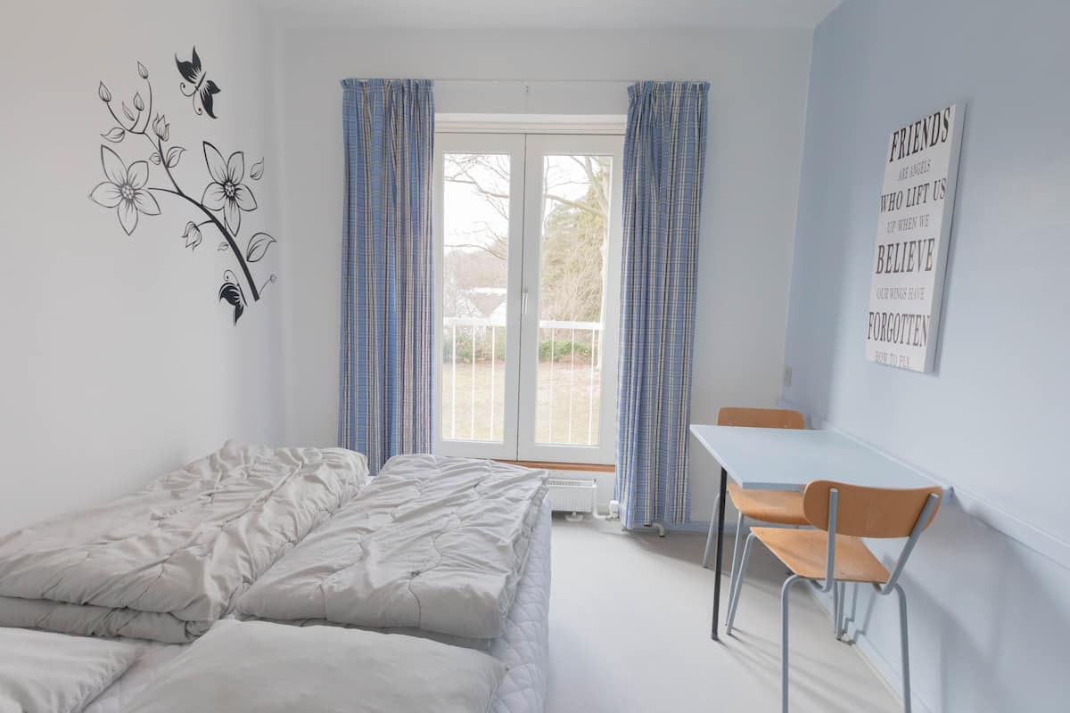 Ein Schlafzimmer mit Bett, Schreibtisch und Stuhl. Es gibt ein Fenster mit Vorhängen, eine Heizung und ein Wandbild.