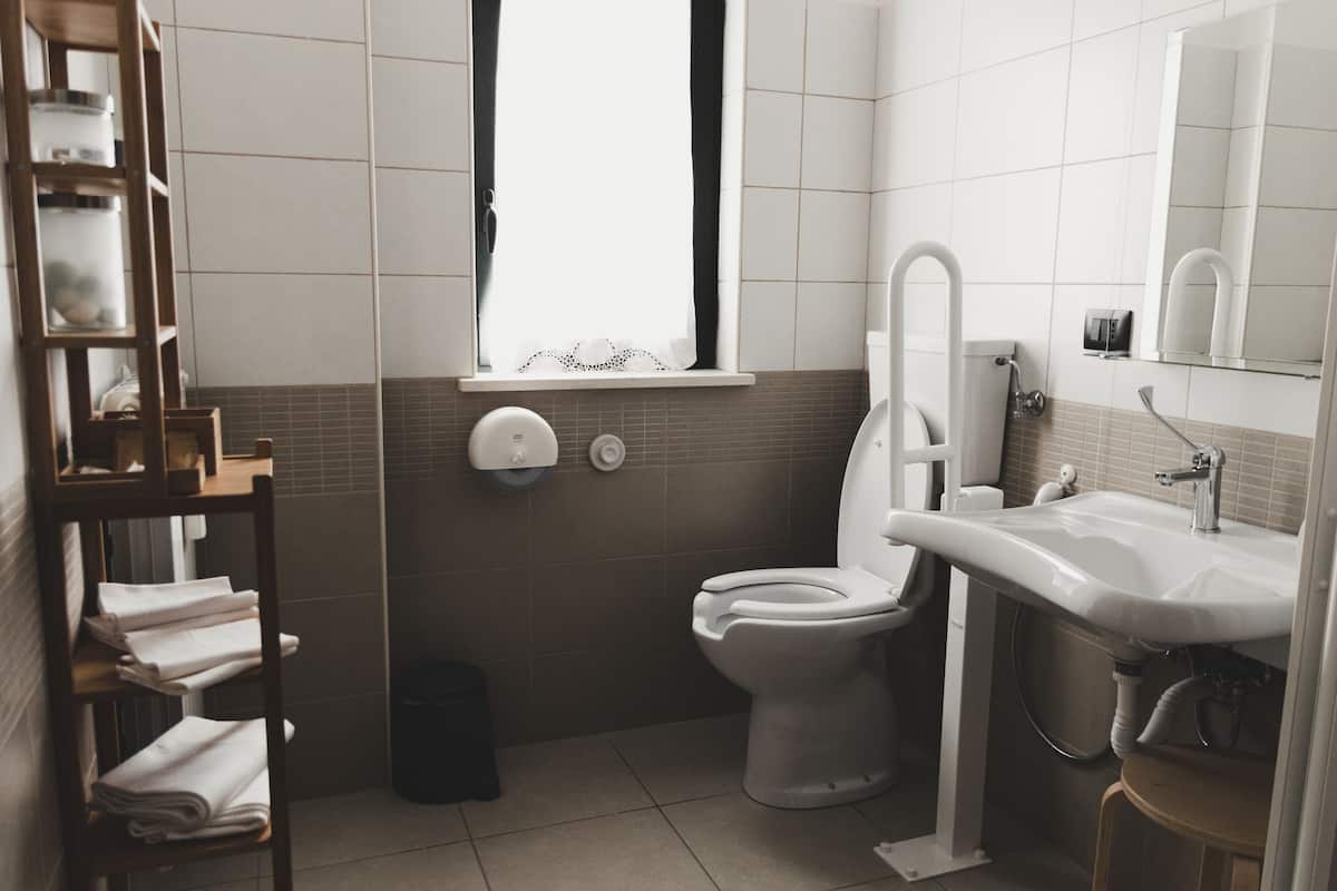 A modern bathroom with a toilet, sink, and a window. There is a shelf with towels and a trash bin.