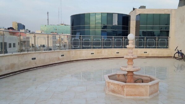 A rooftop terrace with a central fountain, a modern building with reflective glass windows in the background, and a bicycle parked to the side.