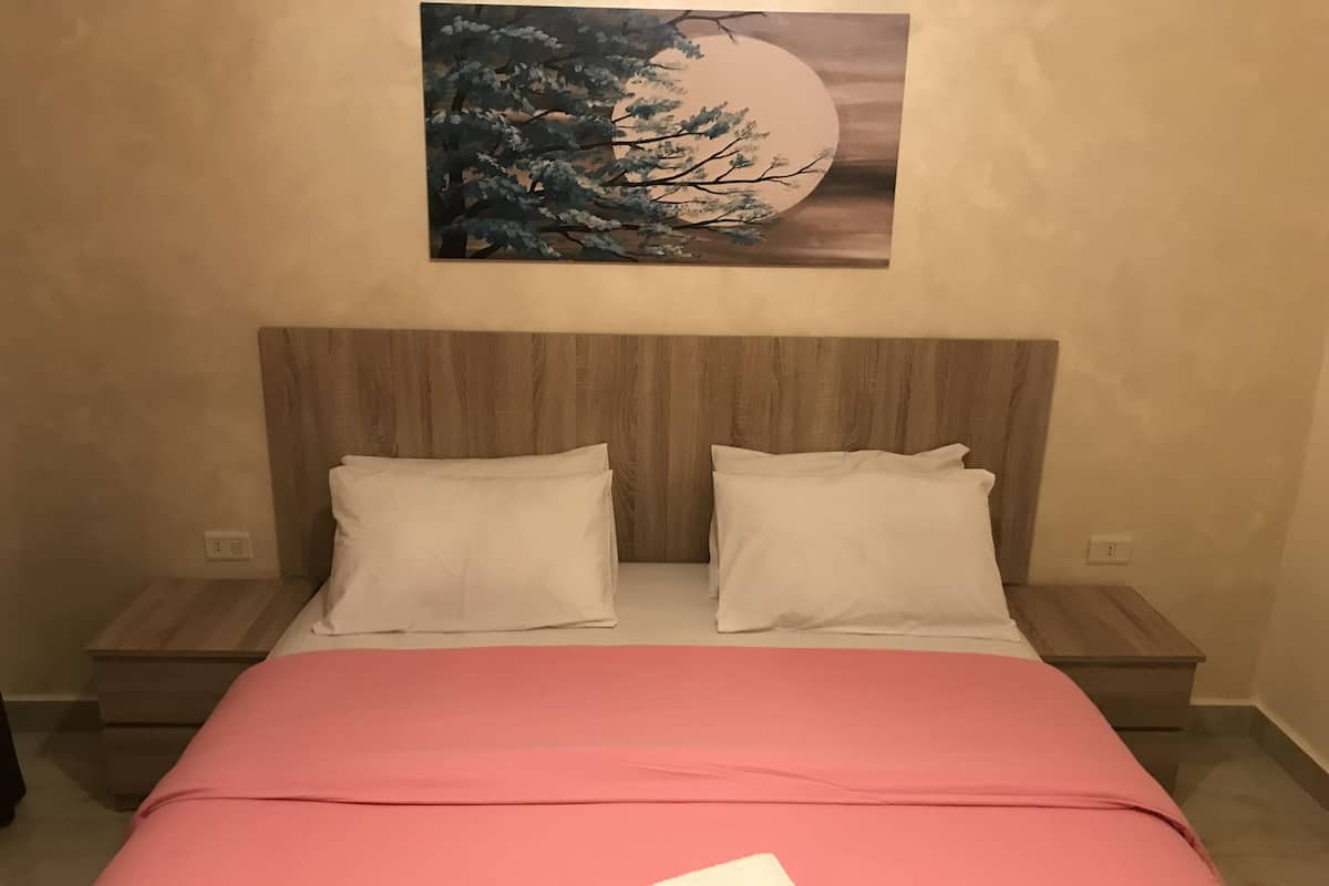 A bed with a pink bedspread, two white pillows, and a painting of a tree and moon on the wall above.