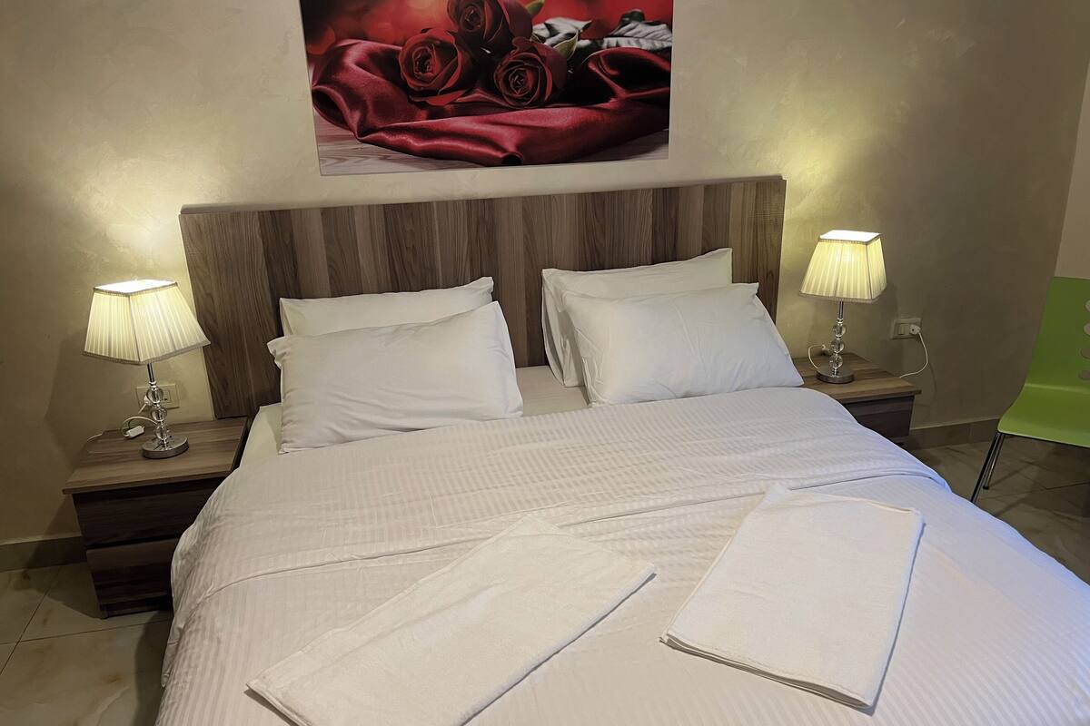 A neatly made bed with white linens, two bedside lamps, and a framed artwork of roses on the wall.