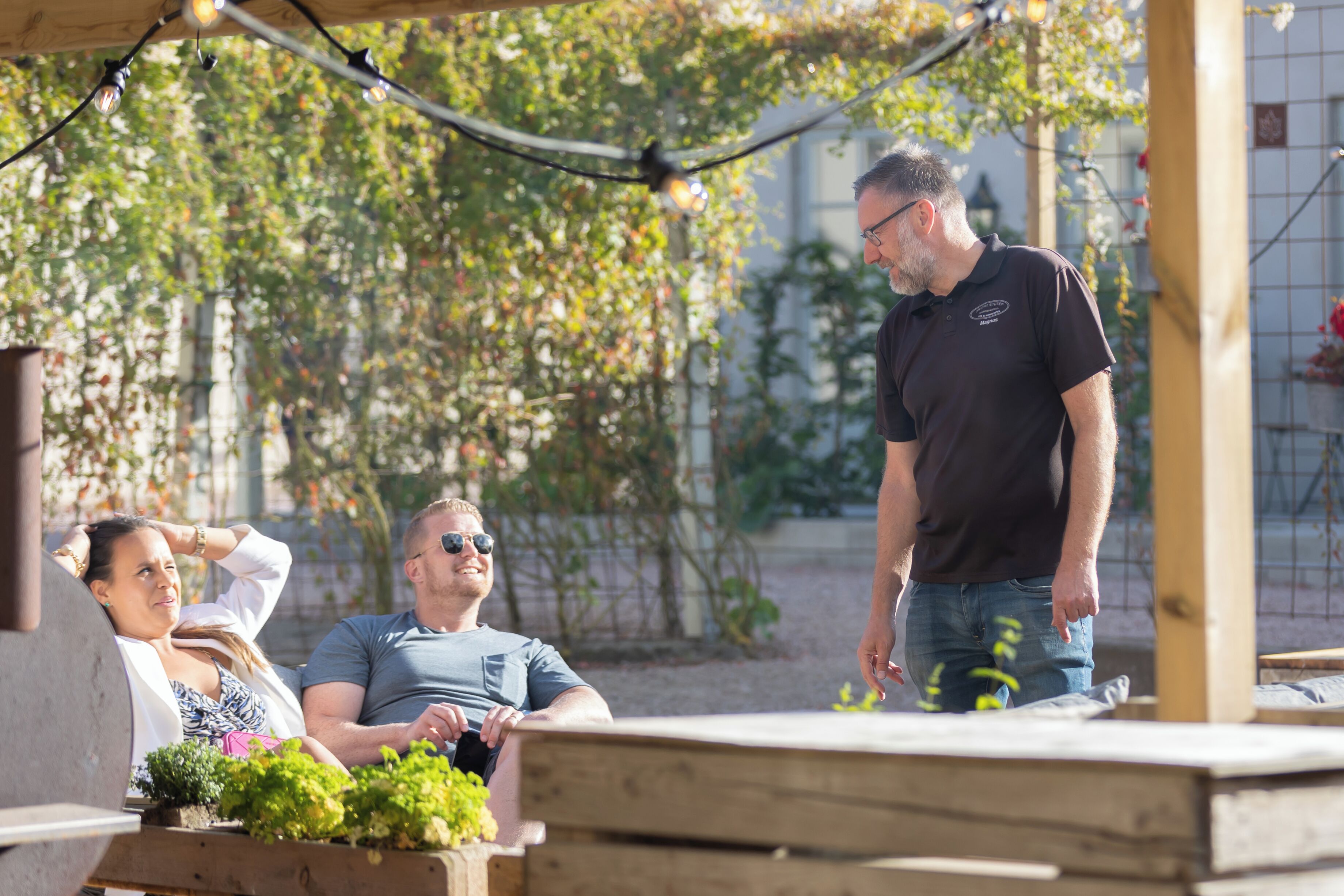 Une terrasse en bois avec une table et des chaises, entourée de verdure et avec un bâtiment en arrière-plan.