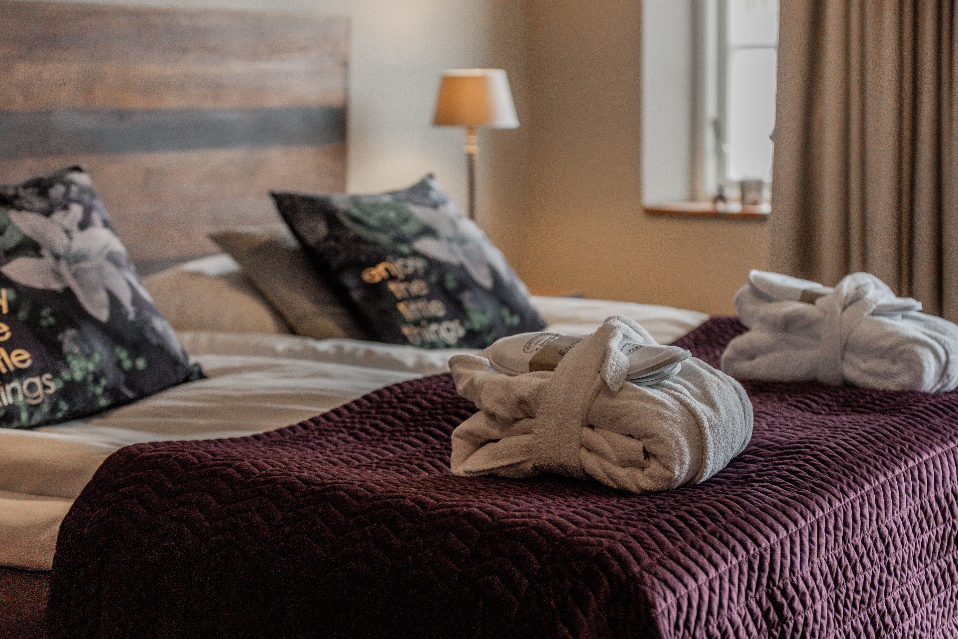 Una cama bien tendida con una colcha rojo oscuro, dos toallas y almohadones con motivos florales.