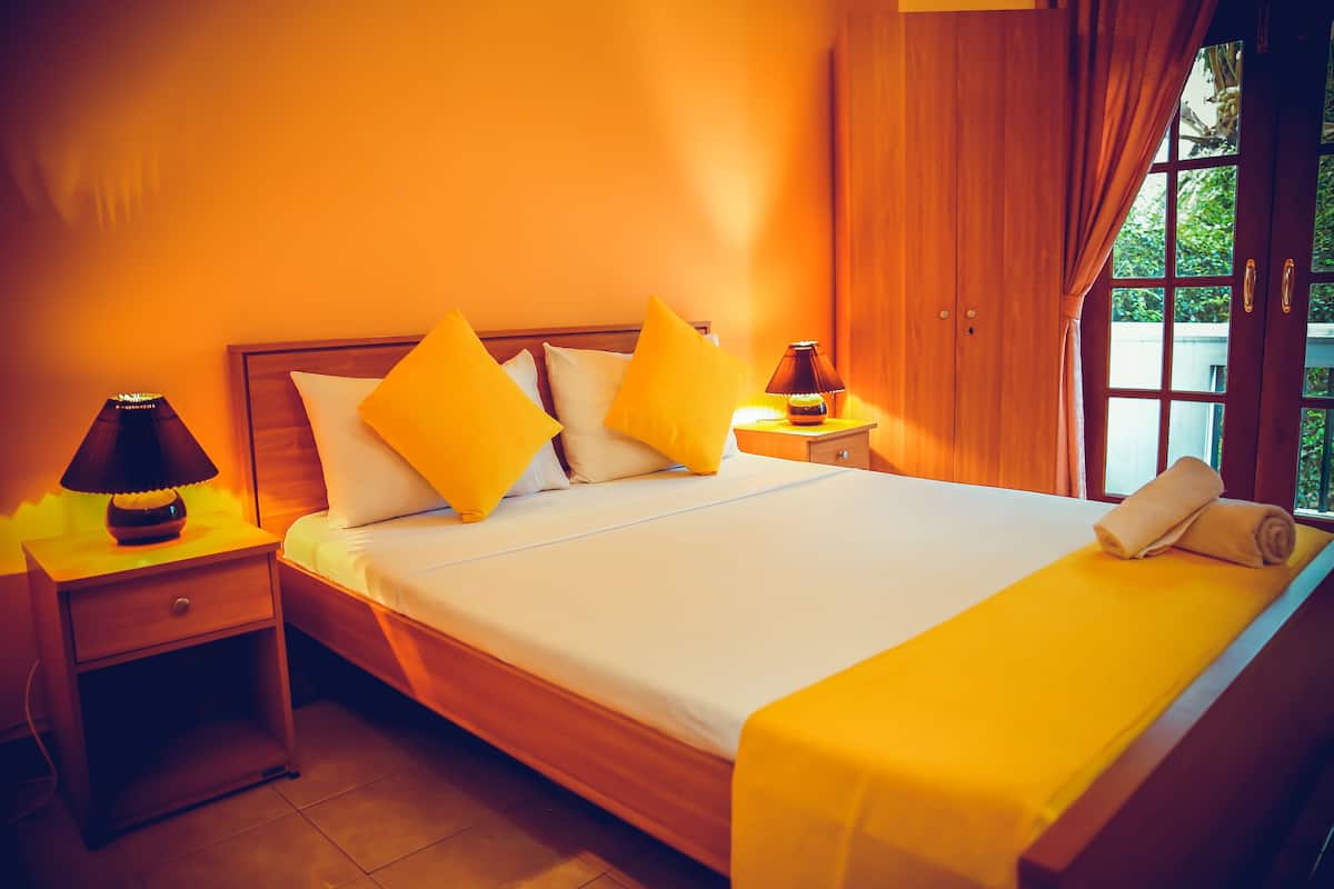 A hotel room with a wooden bed, yellow pillows, a nightstand with a lamp, and a window with curtains.