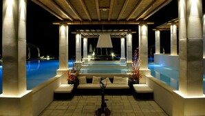 Zona de piscina exterior de noche con una zona de estar cubierta, una fuente central e iluminadas columnas.