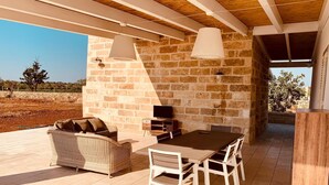 Un patio al aire libre cubierto con pared de piedra, un sofá de mimbre, una mesa de madera con sillas y lámparas colgantes.