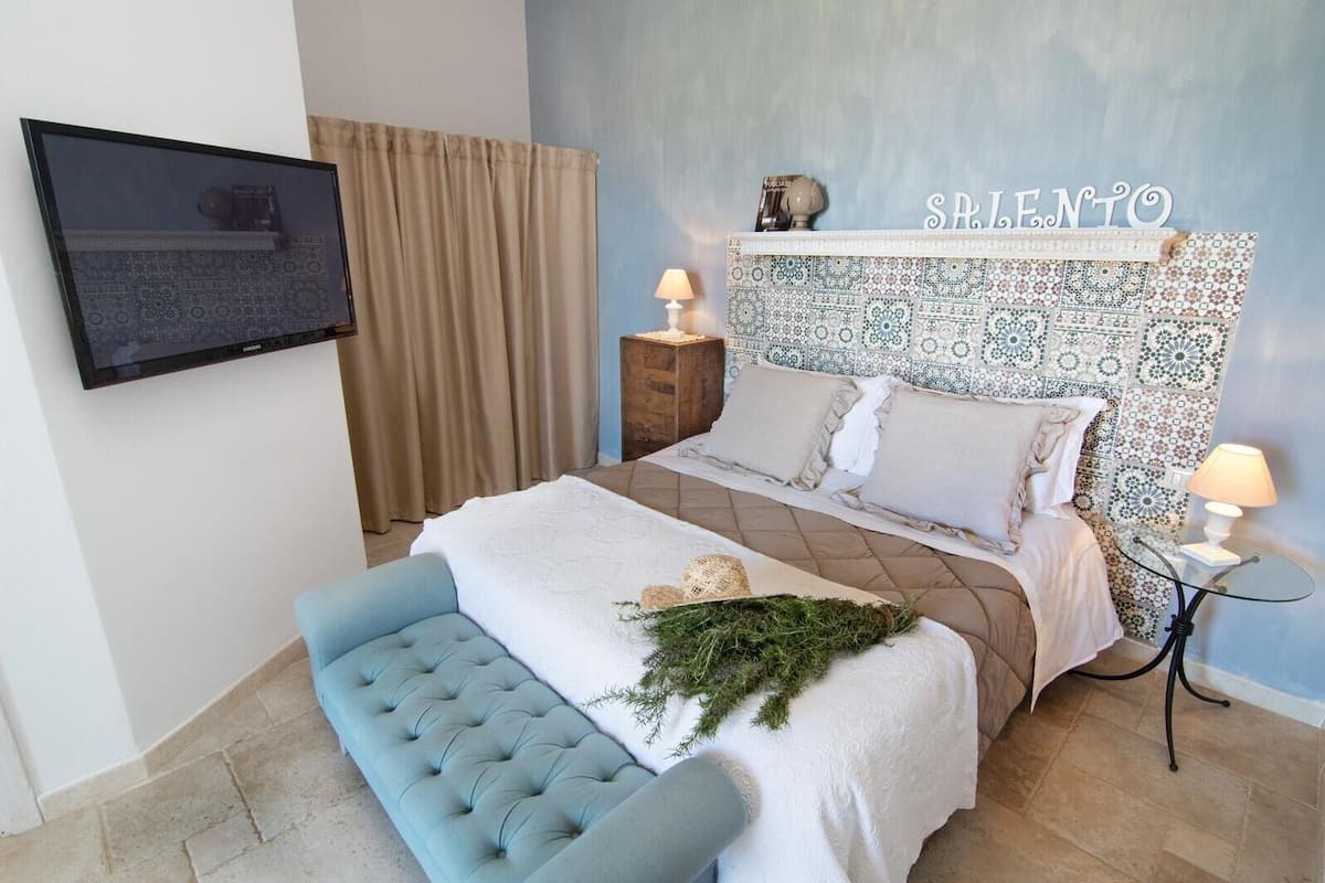 A bedroom with a bed, a blue bench, a wall-mounted TV, a headboard with a nameplate, and a nightstand with a lamp.