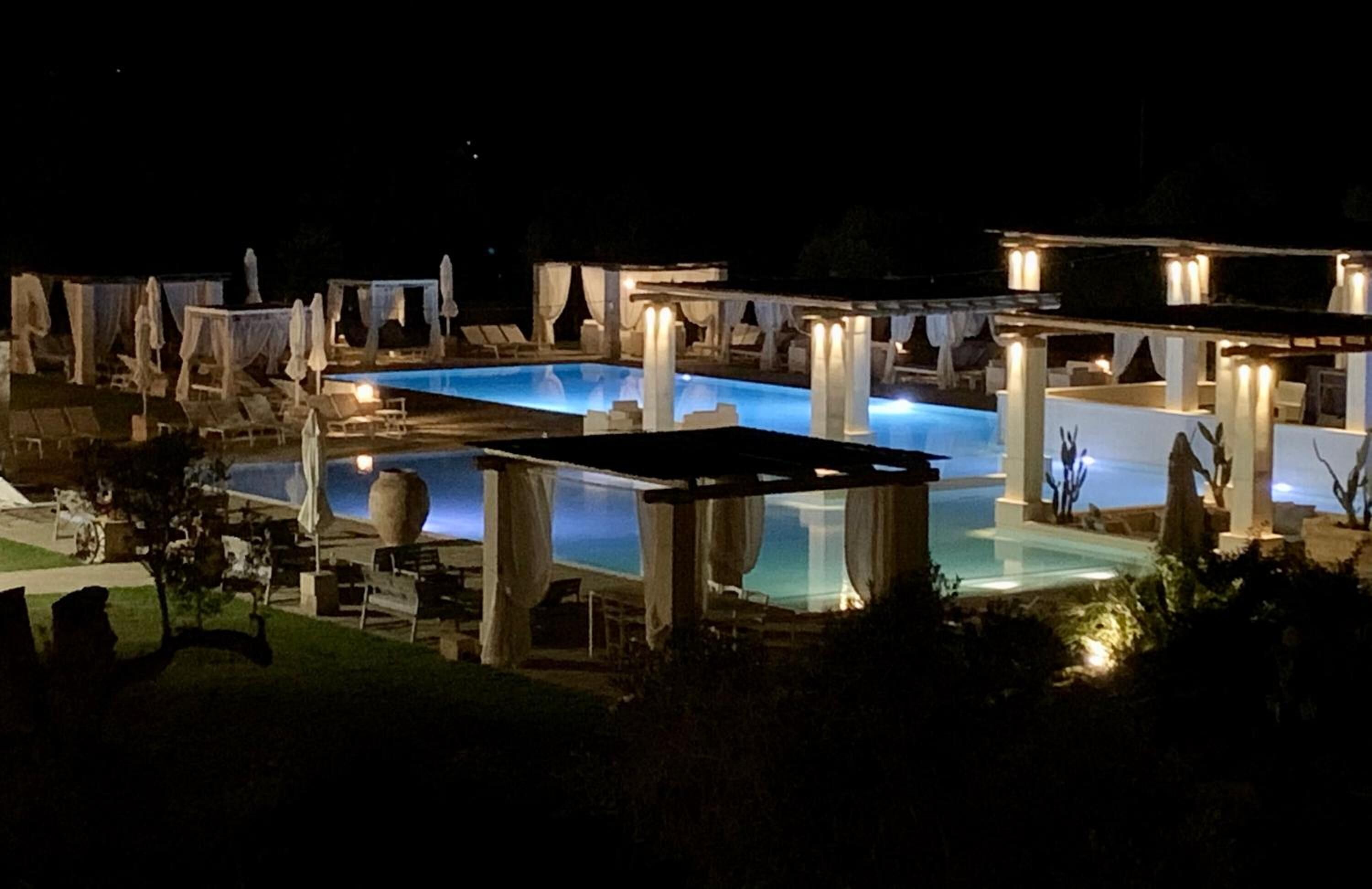 A nighttime view of a pool area with lounge chairs and umbrellas, surrounded by pillars and soft lighting.