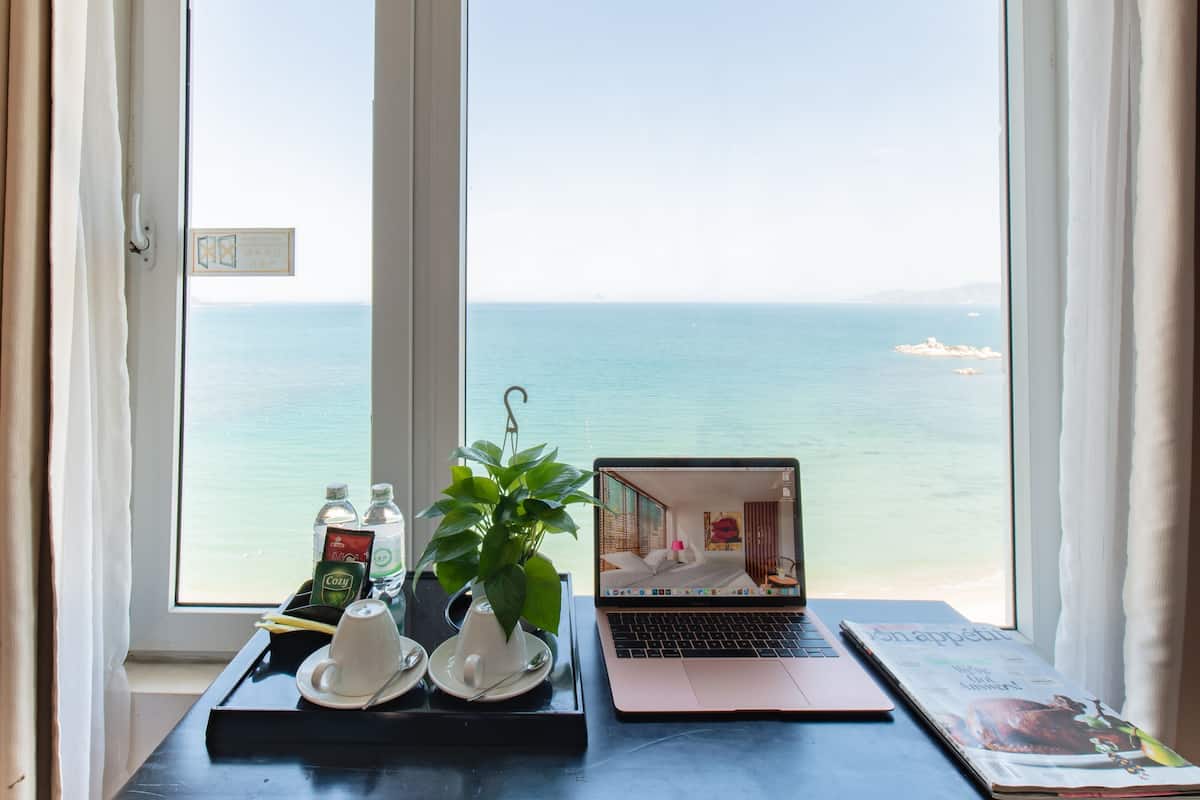 A laptop on a table with a view of the sea through a window.