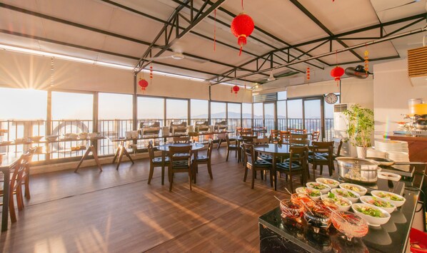 Un restaurante con techos altos, ventanas amplias y vistas a la montaña. Mesas y sillas dispuestas para comer.