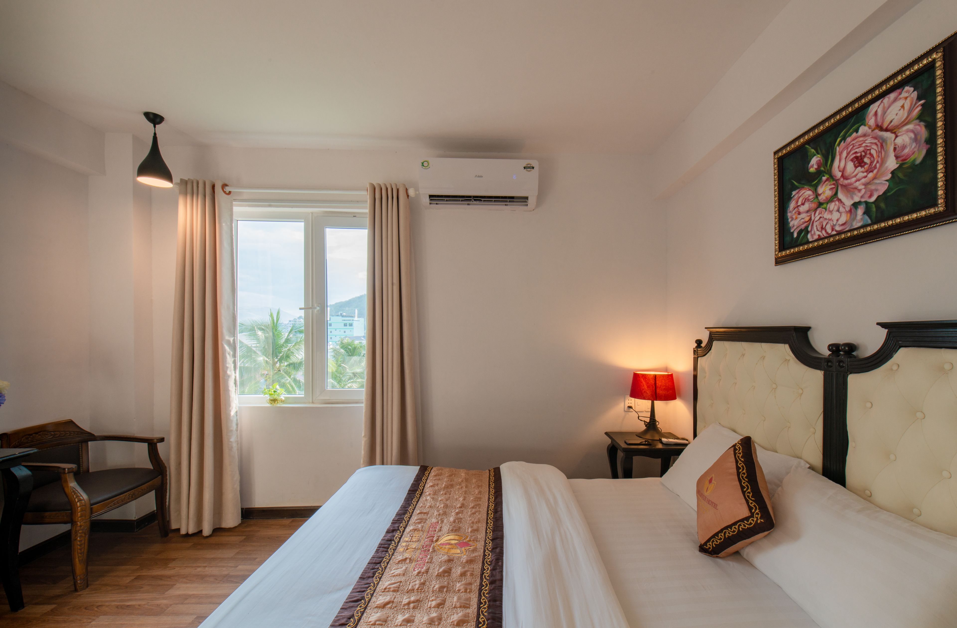 A hotel room with a bed, a desk, a window with curtains, an air conditioner, a lamp, and a painting on the wall.