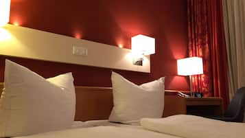 A hotel room with a bed, two white pillows, a wooden headboard, two wall-mounted lamps, a nightstand with a phone and a sign, and red curtains.