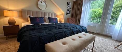 A bedroom with a bed, a bench, a nightstand, a lamp, a mirror, and a window with a view of greenery.