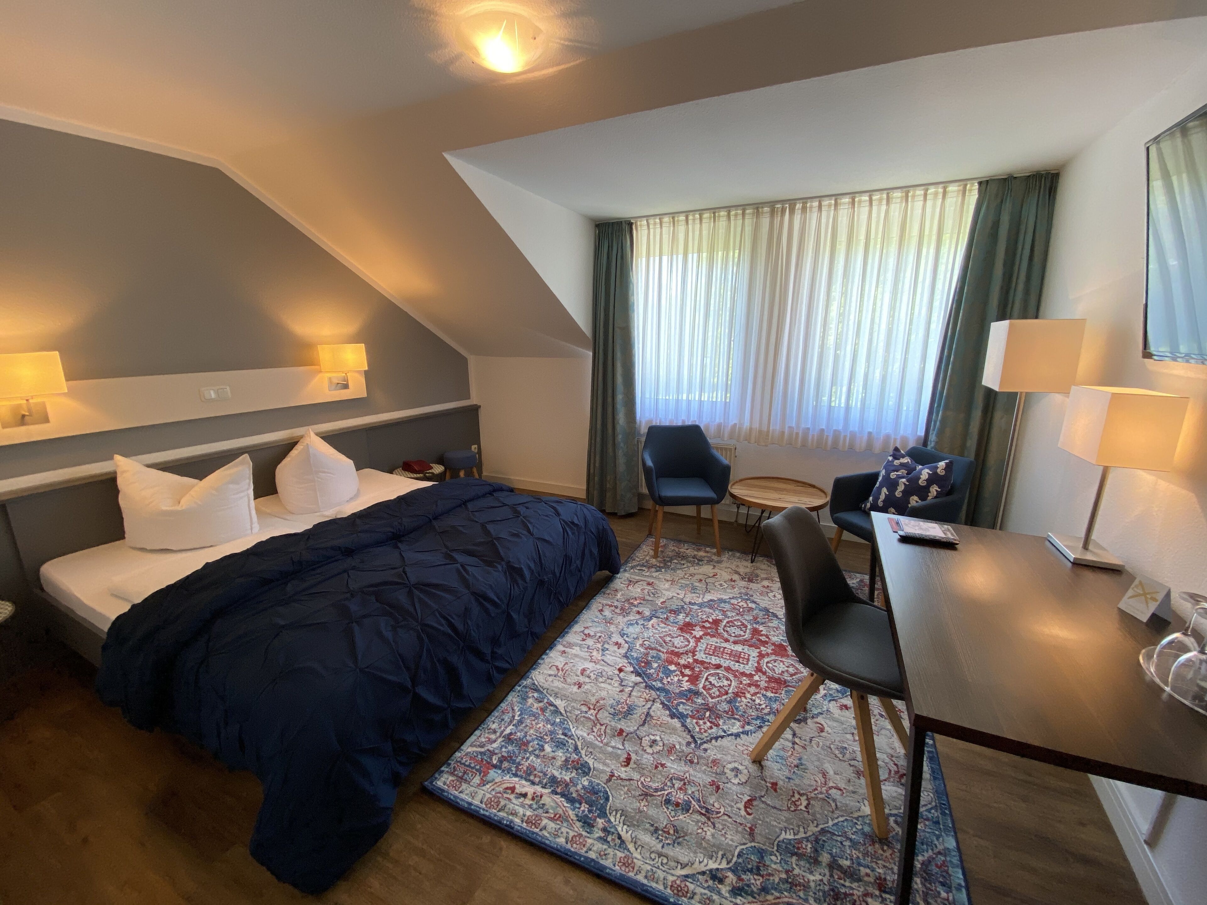 A hotel room with a bed, a desk, a chair, a small table, a lamp, and a window with curtains.