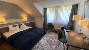 A hotel room with a bed, a desk, a chair, a small table, a lamp, and a window with curtains.