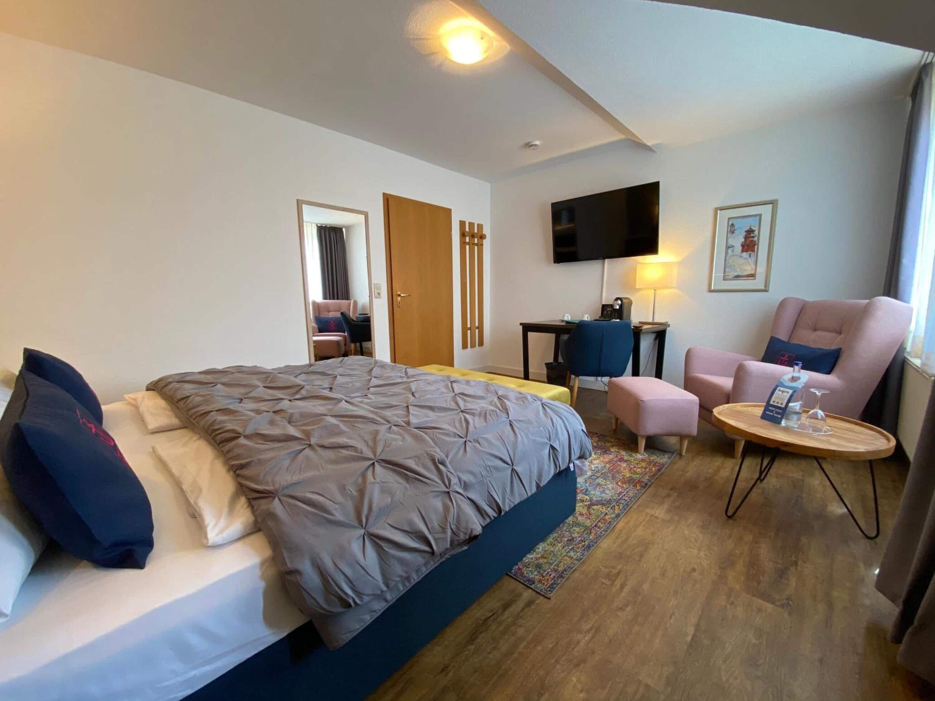 A hotel room with a large bed, a desk, a chair, a TV, and a small table with a bottle and glass.