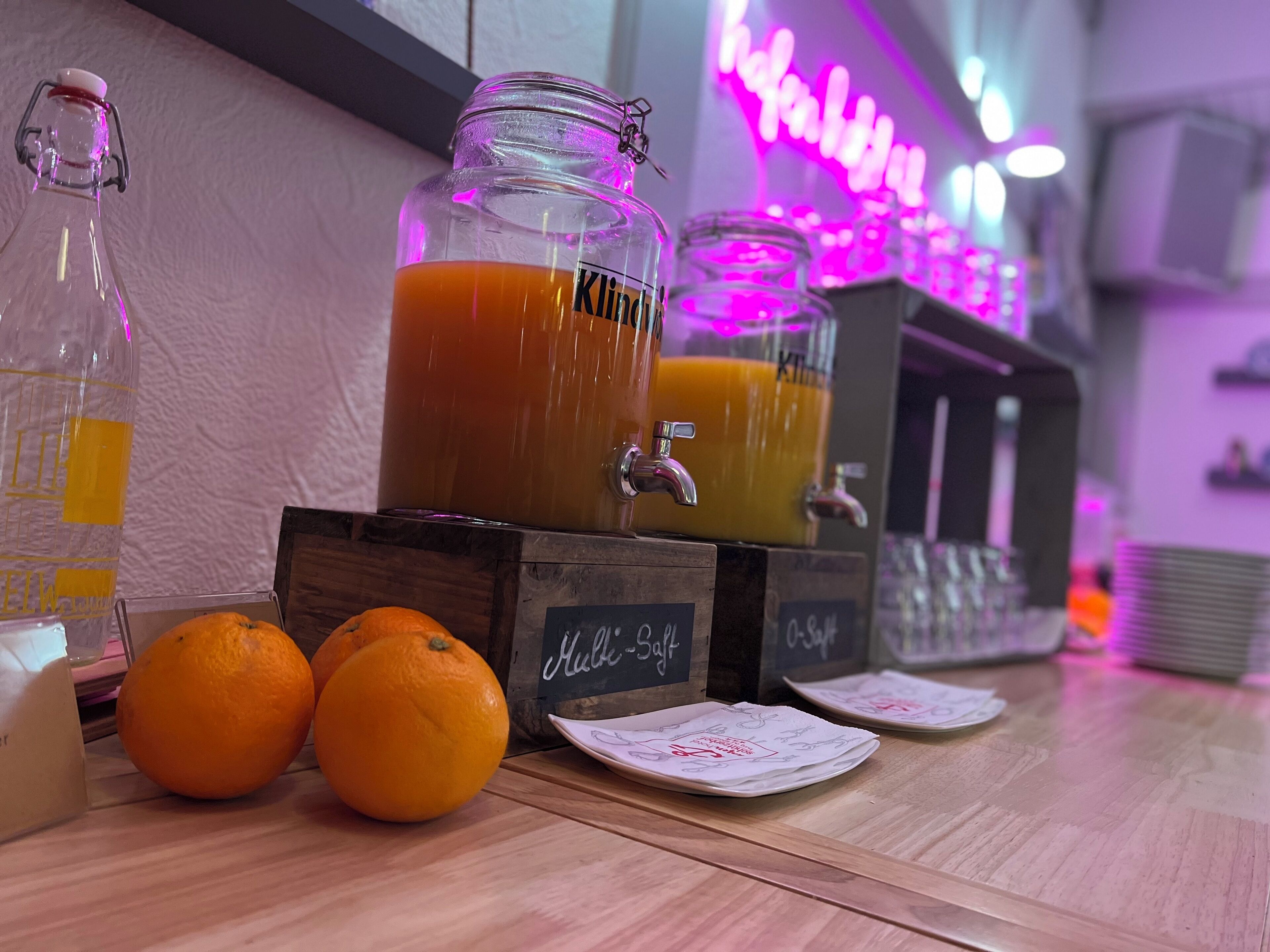 Un coin bar avec un panneau lumineux au néon, un grand bocal en verre rempli de jus d’orange et des oranges disposées sur le comptoir.