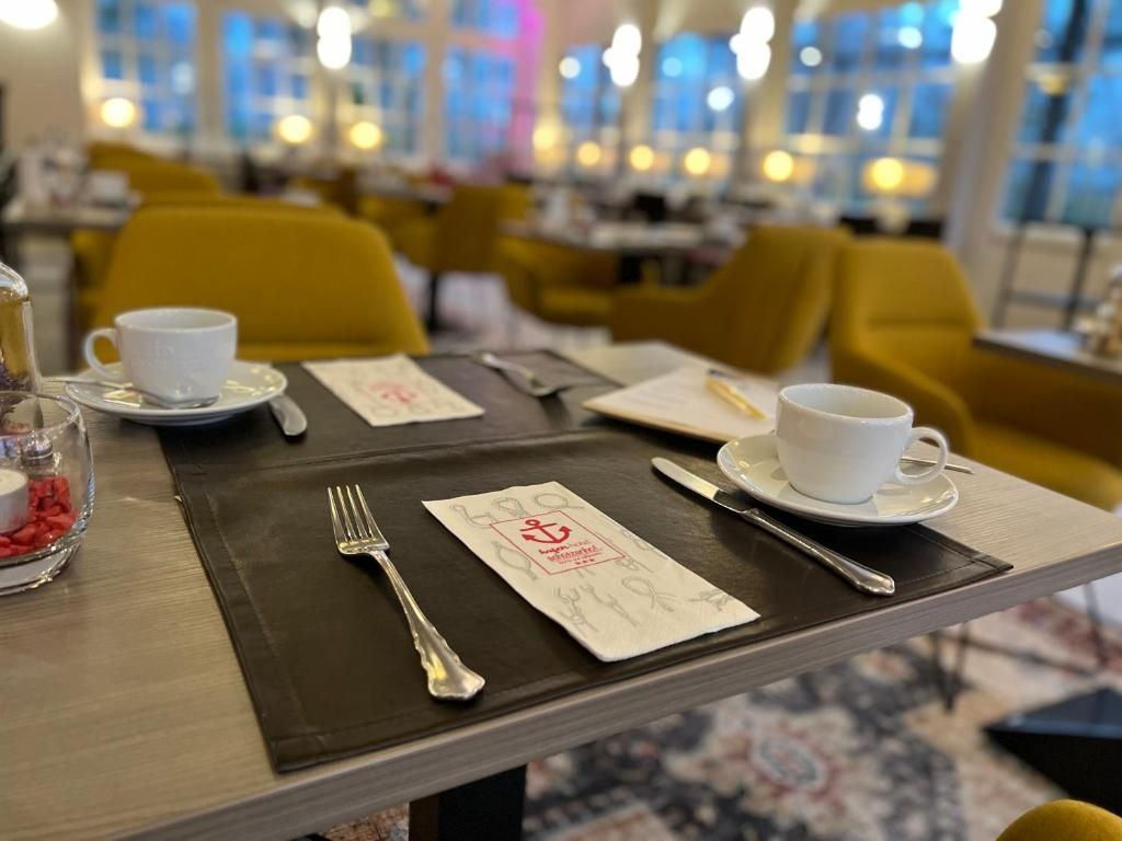 A dining table set with a white cup, saucer, fork, knife, and spoon, along with a menu and a glass of water.