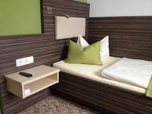Una habitación de hotel compacta con una cama, una mesita de noche, una lámpara y un enchufe.
