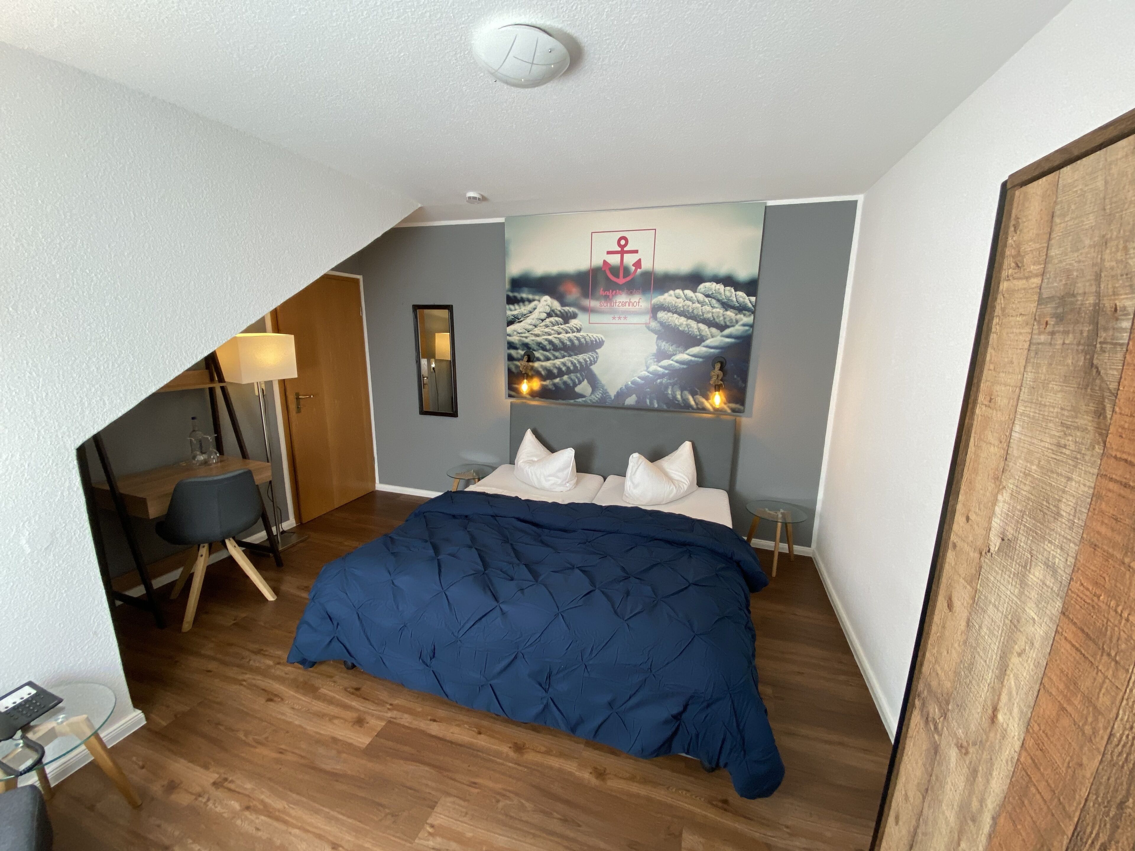A bedroom with a bed, a desk, a chair, a mirror, and a painting of ropes on a wall.