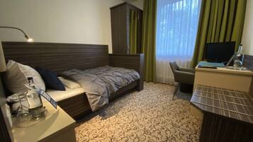 A hotel room with a bed, a desk with a computer, a chair, a wardrobe, and a window with curtains.