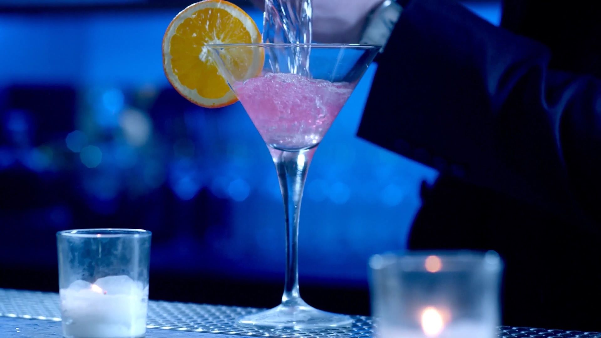 Un vaso de coctel con una bebida rosada, una rodaja de naranja y una vela encendida sobre una mesa.