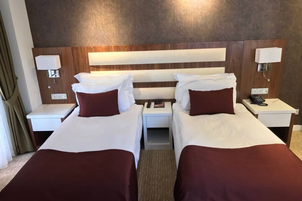 A hotel room with two beds, each with white linens and maroon pillows. There are bedside tables with lamps and a phone.