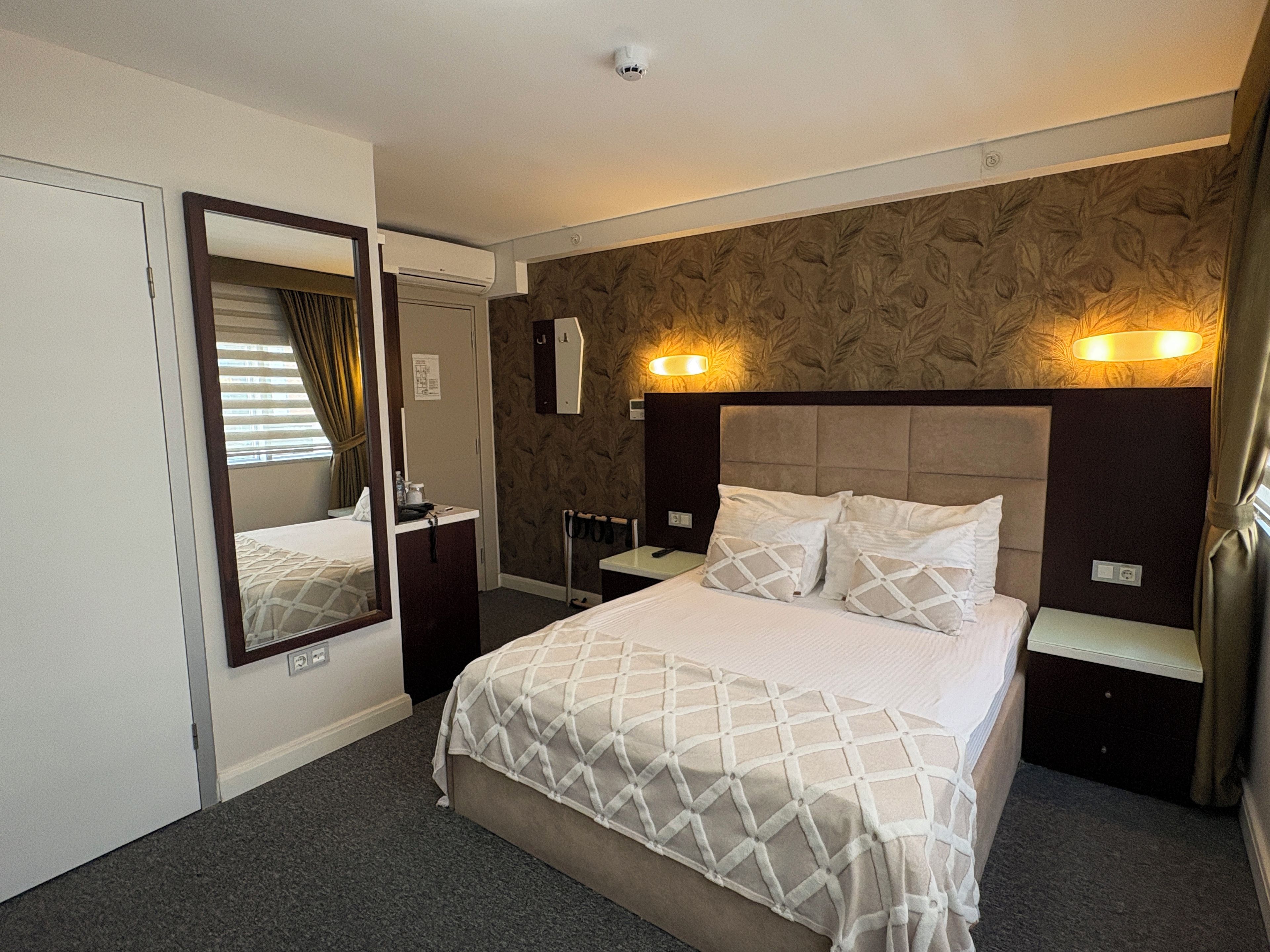 Habitación de hotel con cama, espejo, mesita de noche, luz de pared y ventana con cortinas.