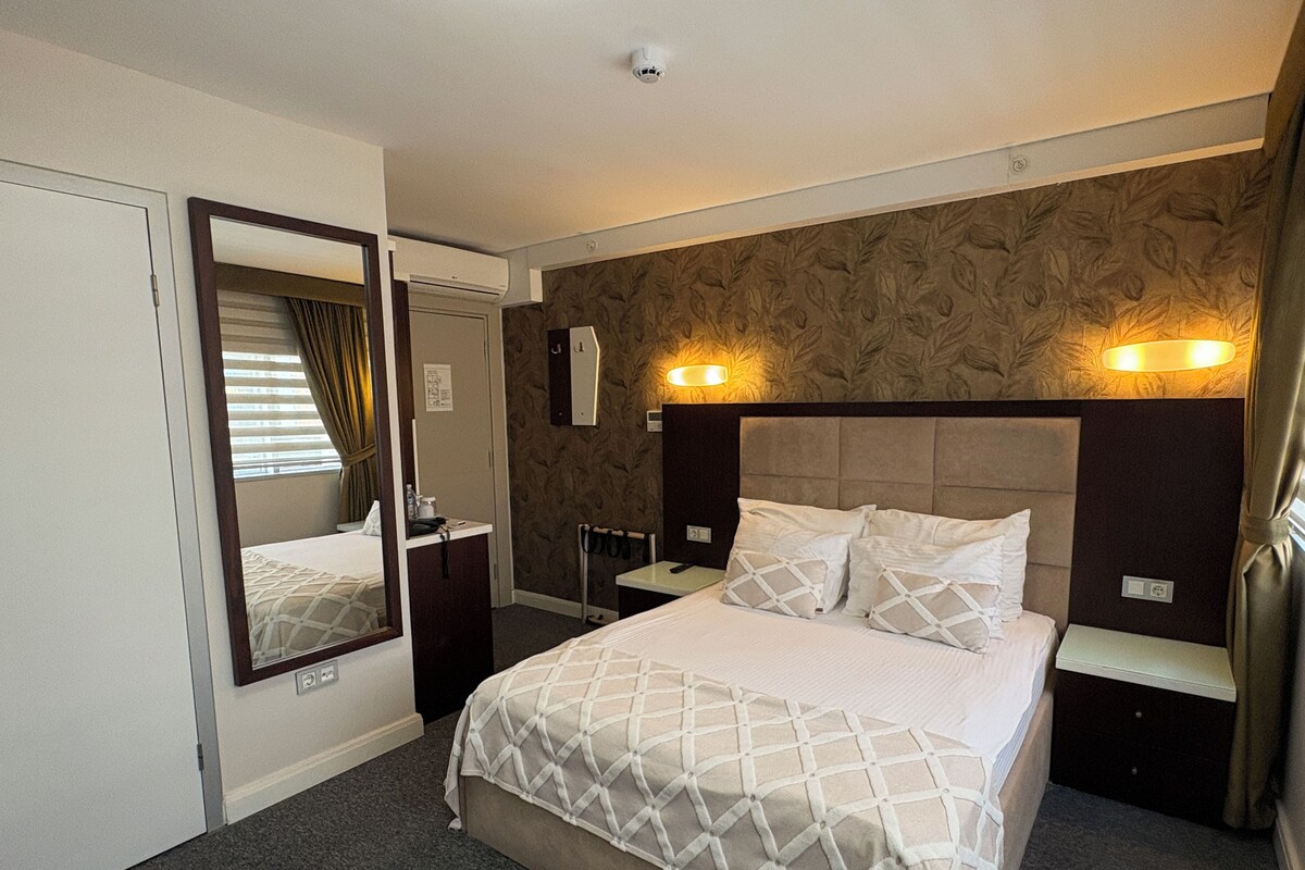 A hotel room with a bed, a mirror, a nightstand, a wall-mounted light, and a window with curtains.