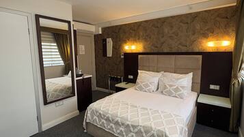 Habitación de hotel con cama, espejo, mesita de noche, luz de pared y ventana con cortinas.