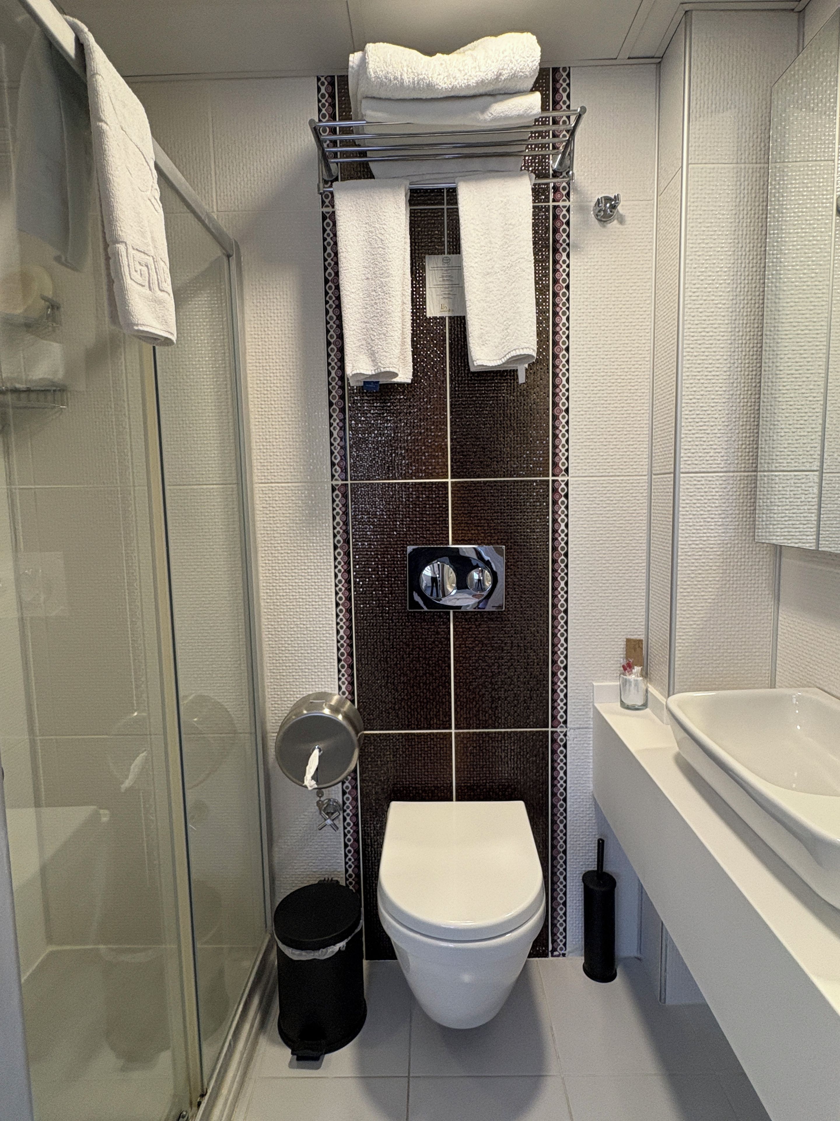 A bathroom with a glass shower enclosure, a toilet, a sink, and a towel rack with folded towels.