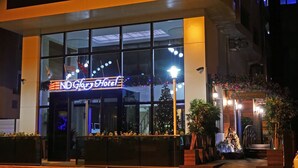 Exterior do Hotel ND Glory com fachada de vidro moderna, sinalização iluminada e área de entrada bem iluminada com plantas em vasos.