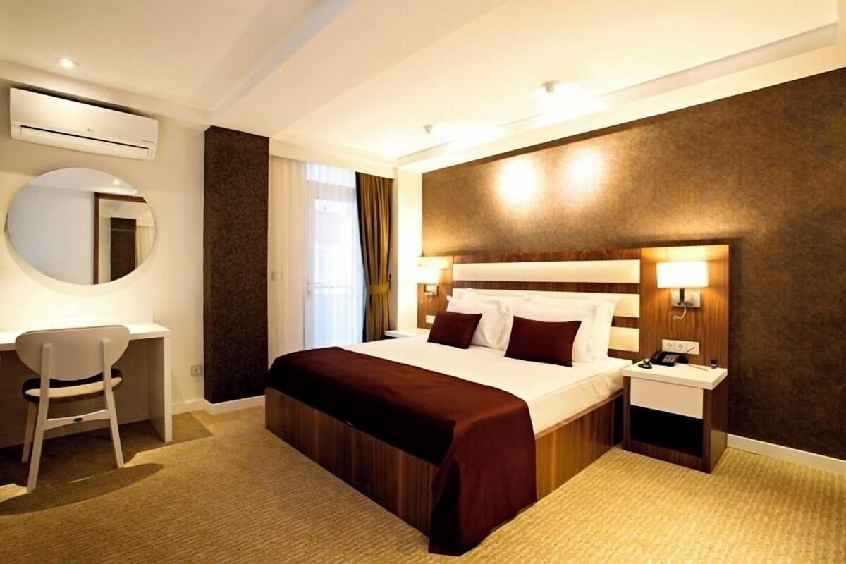 A hotel room with a large bed, a desk, a chair, a round mirror, and a nightstand with a lamp.