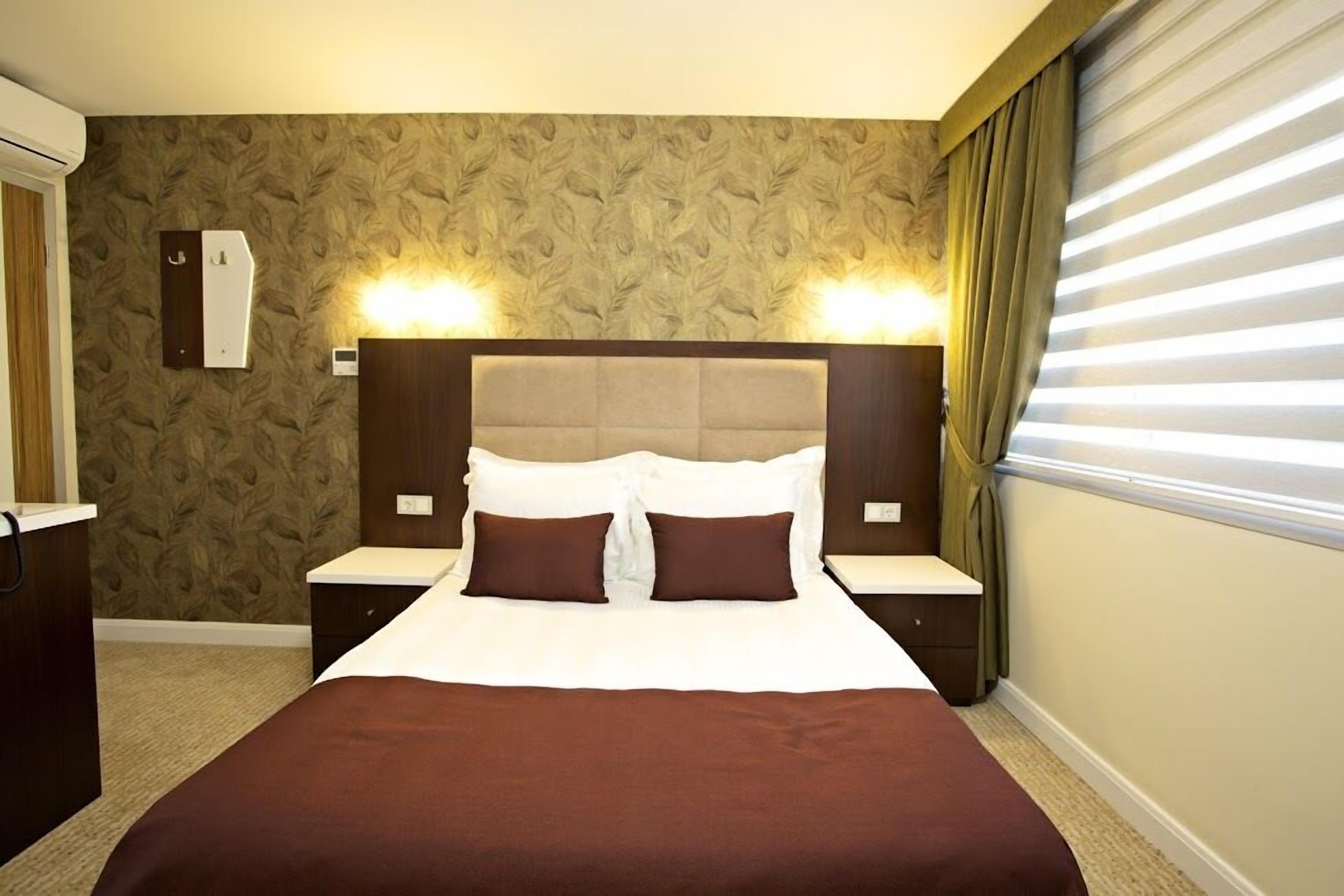 Ein Hotelzimmer mit einem großen Bett, zwei Nachttischen, einem Kopfteil mit integrierter Beleuchtung, einer Wand-Klimaanlage und einem Fenster mit Jalousien.