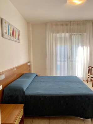 Quarto de hotel com uma cama, mesa de cabeceira de madeira, uma cadeira, um quadro na parede e uma janela com cortinas finas.