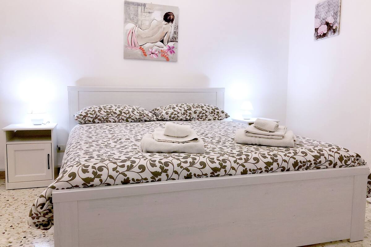 A neatly made bed with patterned bedding, a bedside table with a lamp, and a wall-mounted painting.