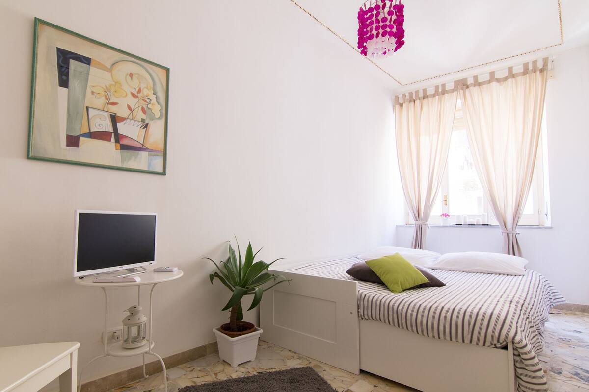 A bedroom with a bed, a desk with a computer, a potted plant, and a painting on the wall.