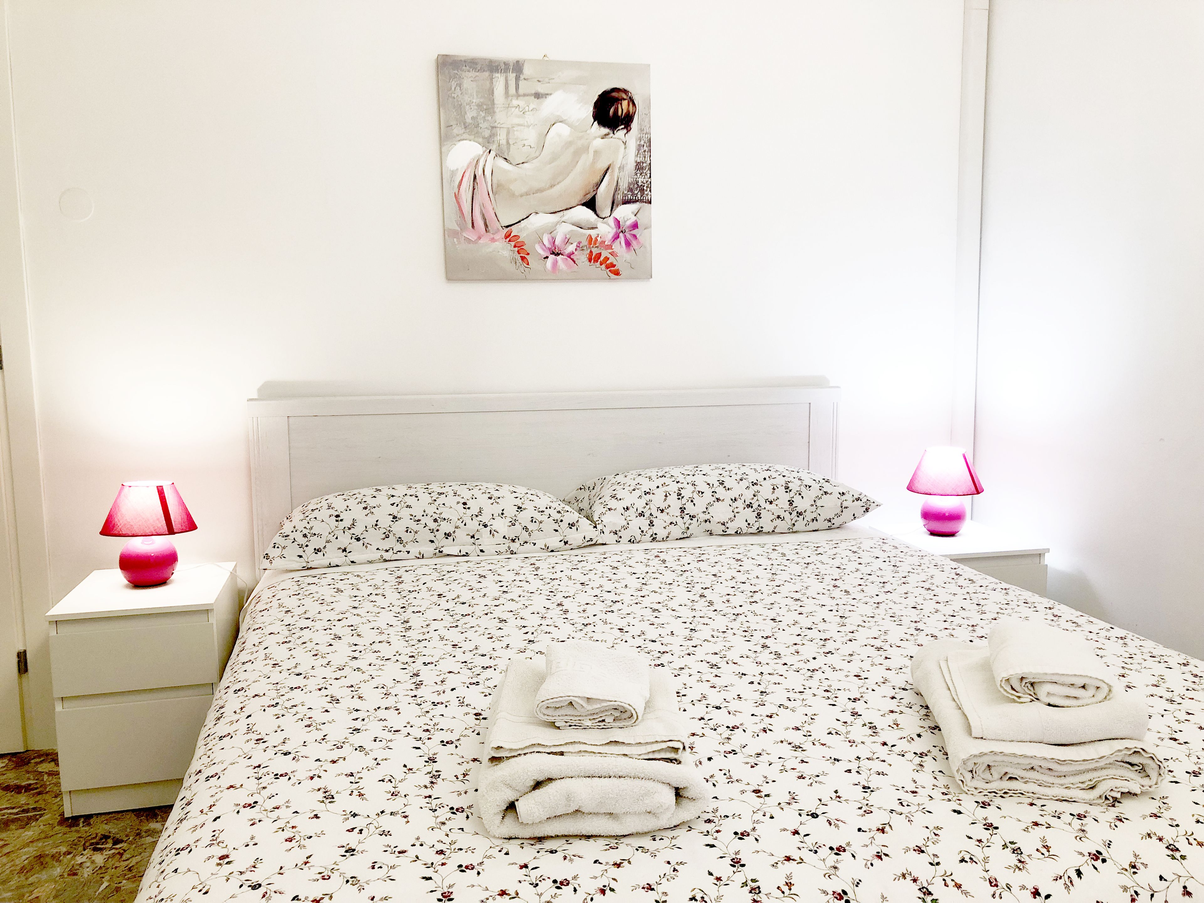 A neatly made bed with floral bedding, two bedside lamps, a painting on the wall, and a stack of towels on the bed.