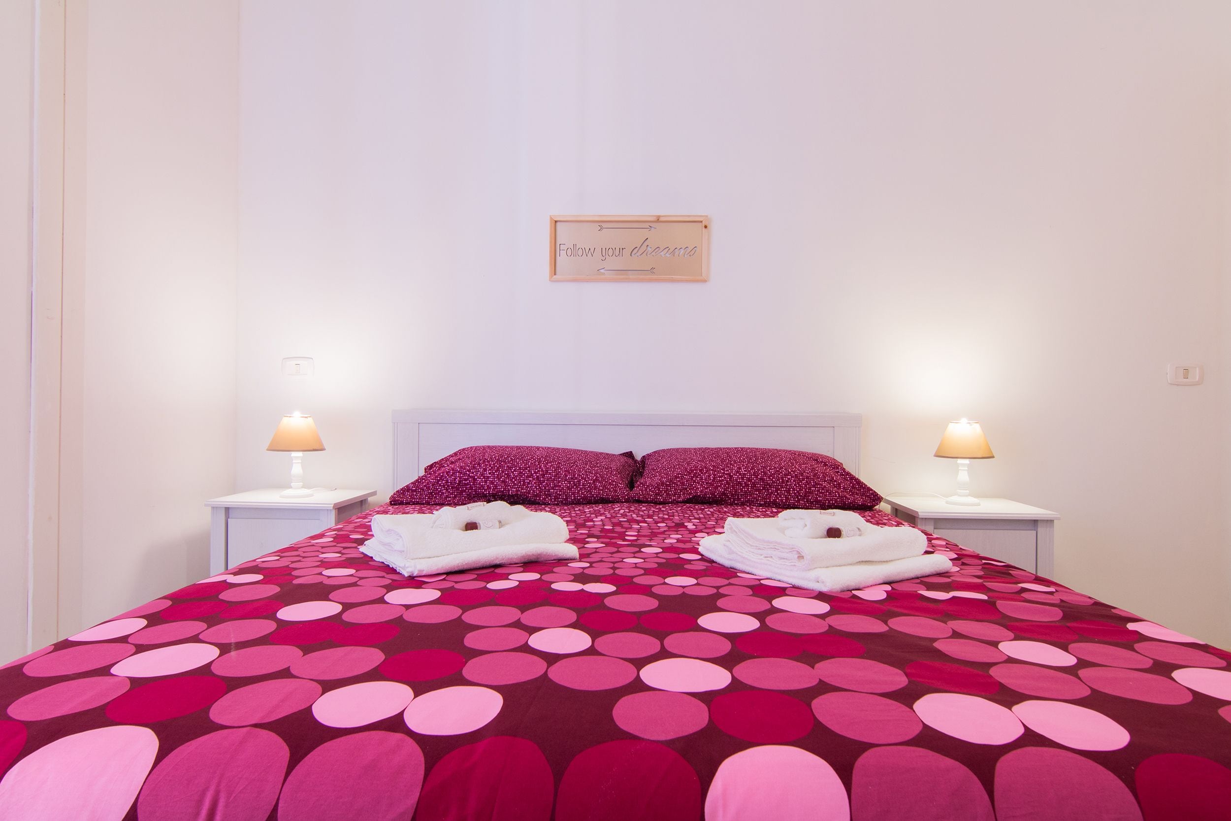 A neatly made bed with a polka dot bedspread, two pillows, and folded towels on either side. There is a sign above the bed and two bedside lamps.