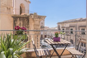 A balcony with a wooden table and chairs, potted plants, and a view of historic buildings. - Maison Ortigia (Syracuse)