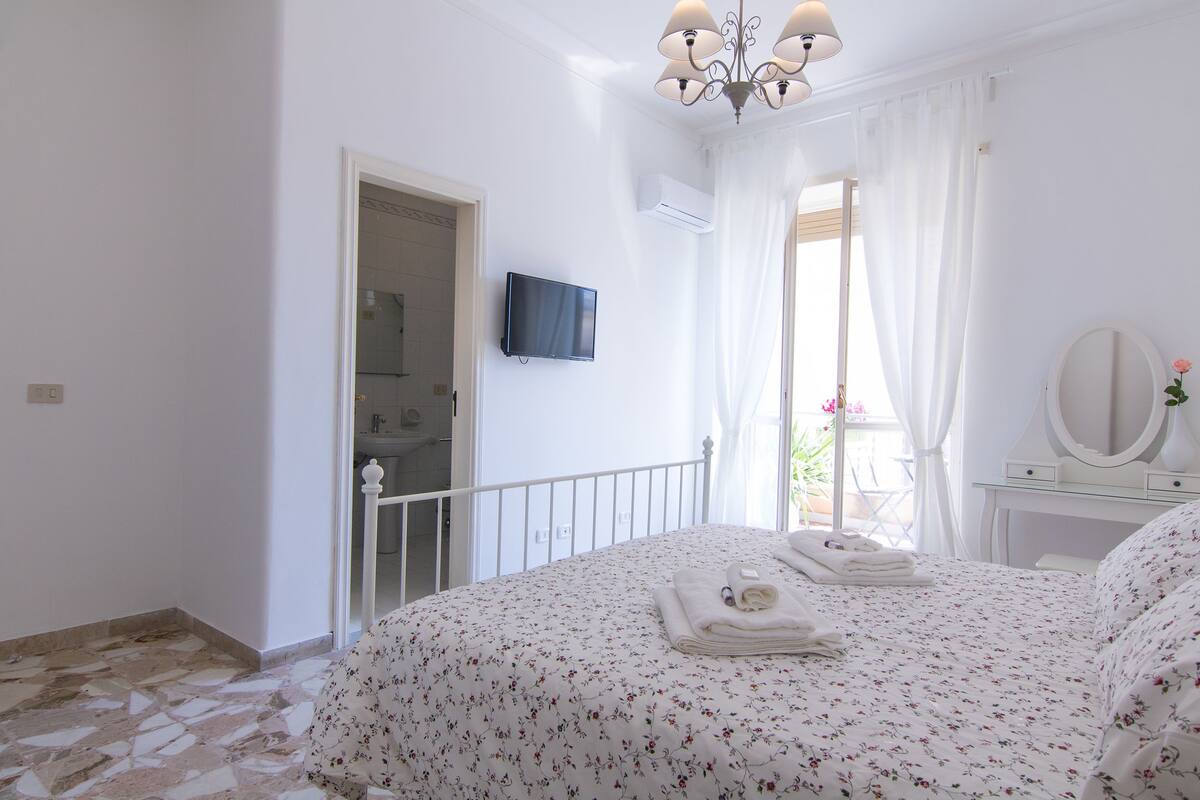 A bedroom with a bed, a TV, a mirror, and a balcony with a view.