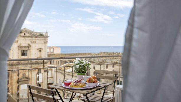 A balcony with a table set for breakfast, a potted plant, and a view of the sea and historic buildings.