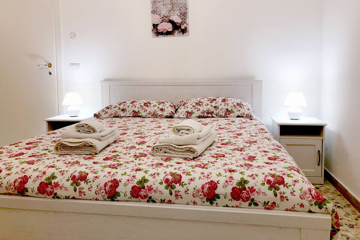 A neatly made bed with floral bedding, two bedside lamps, and a wall-mounted picture.