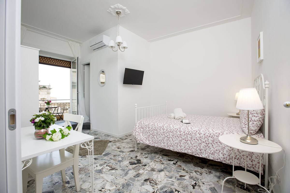 A bedroom with a bed, a bedside table, a chair, a TV, and a window with curtains.