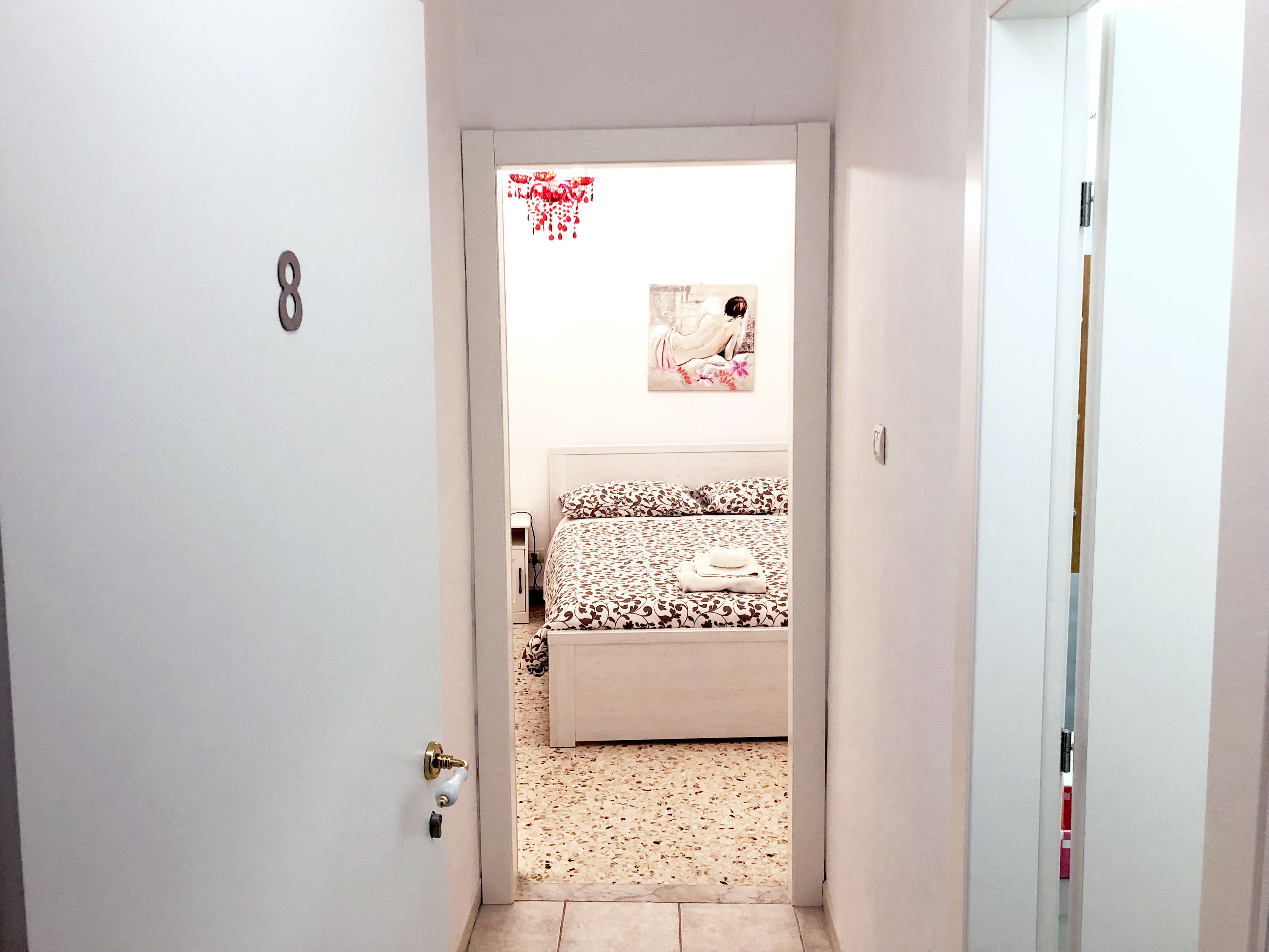 A narrow hallway leading to a bedroom with a bed, a wall-mounted picture, and a door with a number plate.