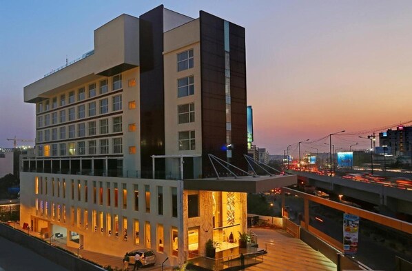 Un hotel moderno con varios pisos, una terraza en la azotea y vista del paisaje urbano al atardecer.