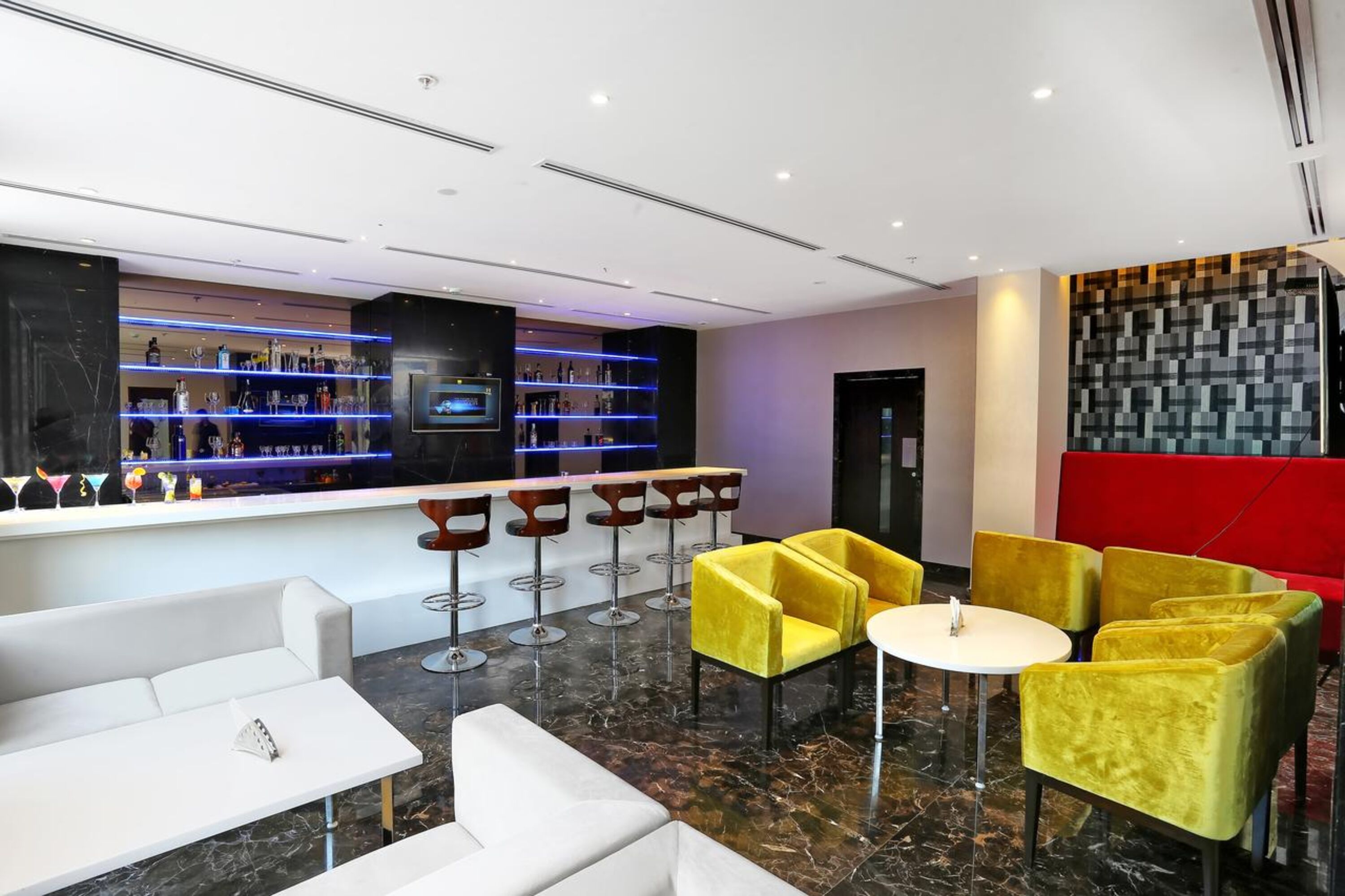 A modern bar area with a marble floor, yellow chairs, white sofas, and a red accent wall.