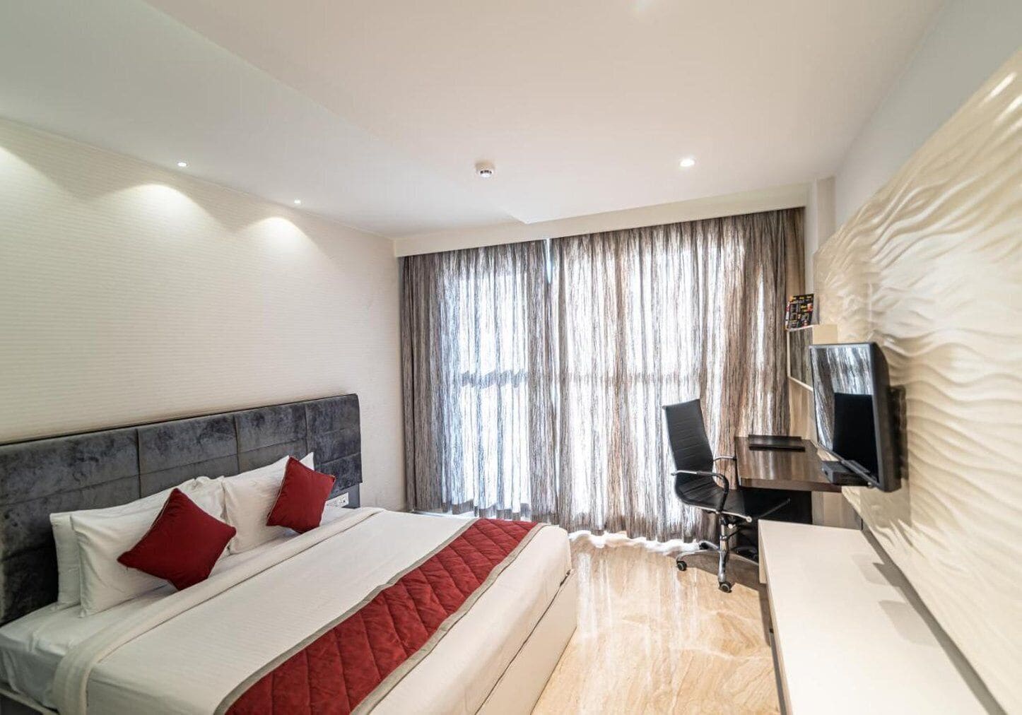 Ein modernes Hotelzimmer mit einem großen Bett, einem Schreibtisch mit Stuhl, einem flachbildfernseher an der Wand und einem großen Fenster mit Vorhängen.