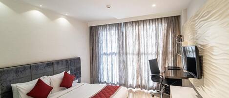 A modern hotel room with a large bed, a desk with a chair, a flat-screen TV mounted on the wall, and a large window with curtains.