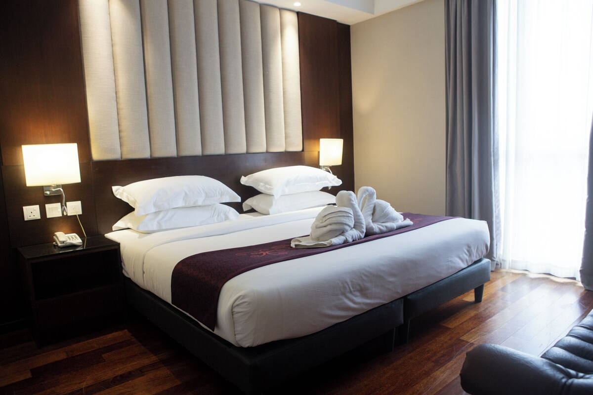 Una habitación de hotel con una cama grande, dos mesitas de noche, un buró, piso de madera oscura y una ventana con cortinas.